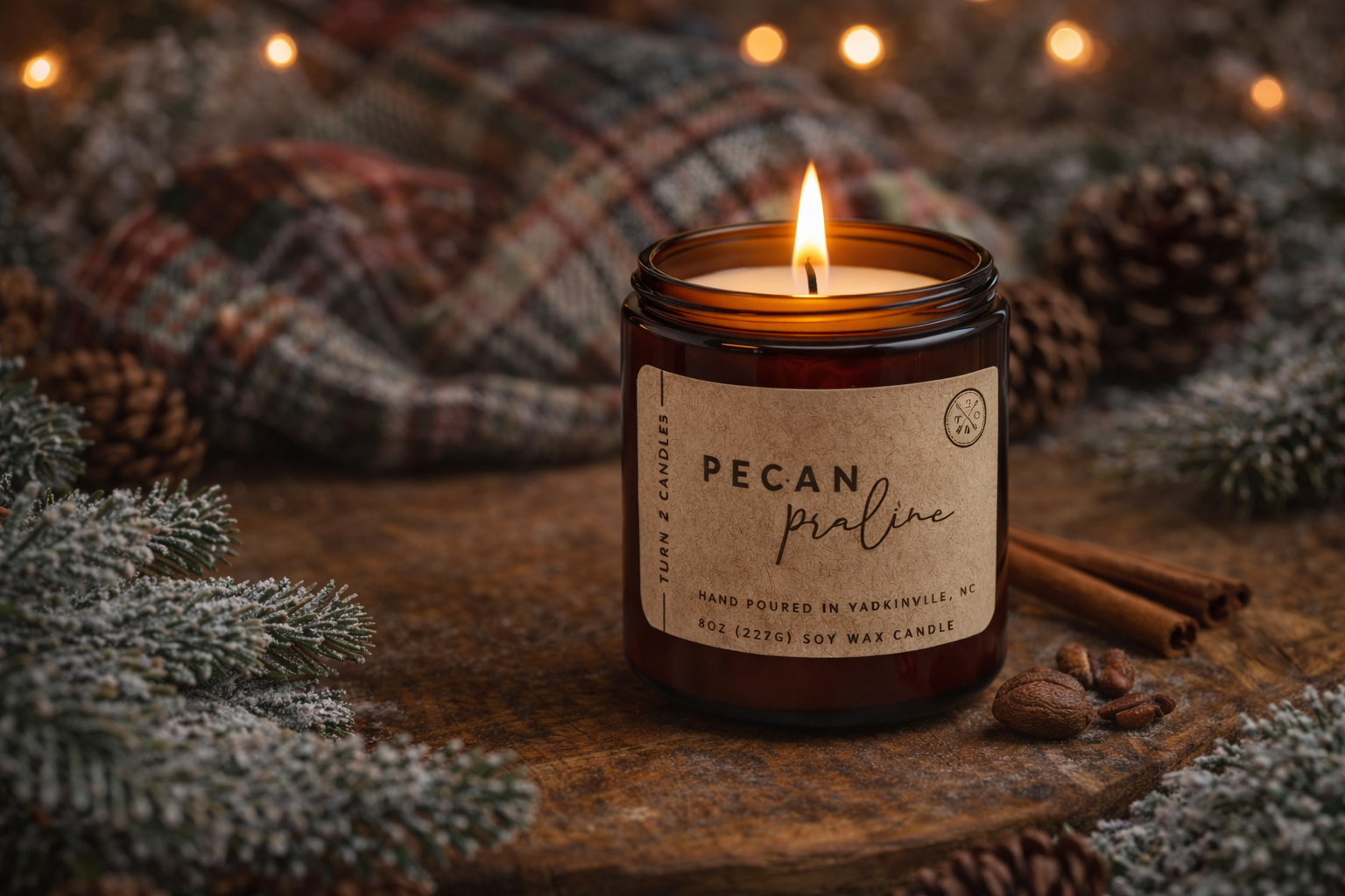 lit amber glass soy candle labeled "Pecan Praline" resting on a rustic wooden surface surrounded by frosted evergreen branches, pinecones, cinnamon sticks, and pecans, creating a warm winter candle scene