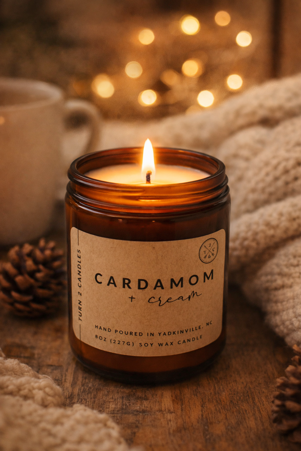 An amber glass soy candle labeled ‘Cardamom + Cream’ burns with a warm flame on a wooden surface, surrounded by a cozy knit blanket, soft fairy lights, and a ceramic mug, creating a warm, inviting atmosphere.