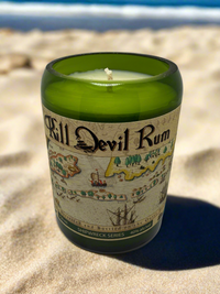 Thumbnail for A green upcycled alcohol bottle candle with 'Kill Devil Rum' text on it, placed on a sandy surface with a blue sky in the background.