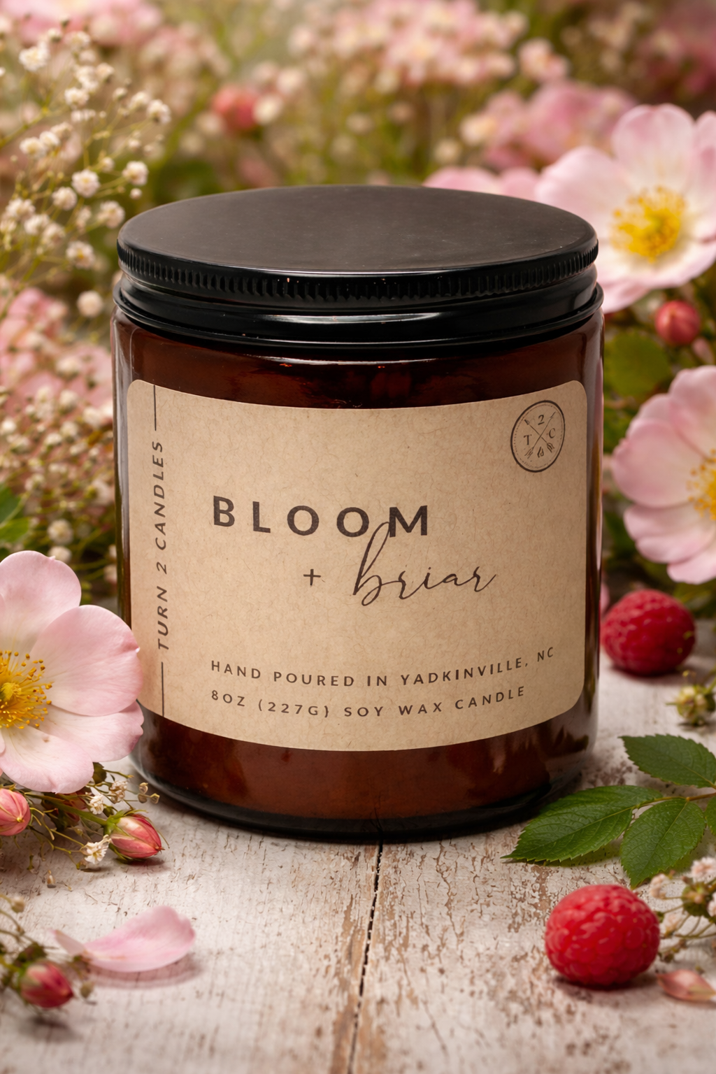 Candle jar with 'Bloom + Briar' label surrounded by flowers and berries on a wooden surface
